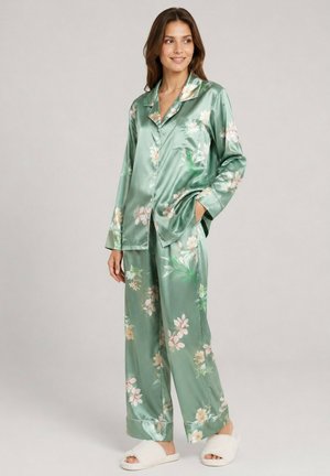 Femme debout portant un ensemble pyjama en satin vert à fleurs, manches longues, avec pantalon assorti et chaussons blancs duveteux sur fond neutre.