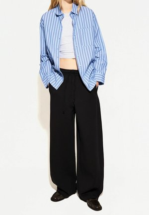 Person trägt ein blau gestreiftes Hemd mit Knöpfen über einem weißen Crop-Top, weite schwarze Hosen mit Kordelzug und schwarze Schuhe und steht vor einem schlichten Hintergrund.