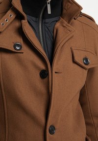 Abrigo de mezcla de lana marrón con superficie texturizada, botones negros, capa interior con cremallera y epaulettes decorativos en los hombros.