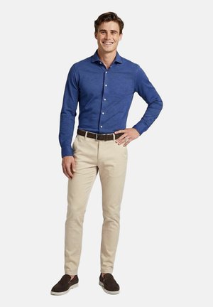 Lächelnder Mann steht mit einer Hand in der Hüfte, trägt ein blaues Hemd mit Knöpfen, beige Hose, dunklen Gürtel und braune Slipper vor weißem Hintergrund.