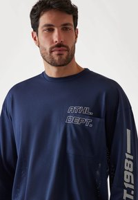 Donkerblauwe lange mouwen mesh sportshirt met een voorzak, de tekst "ATHL. DEPT." en zijaccentdetails in lichtgrijs.