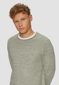 Hellgrüner, gestrickter Pullover mit strukturierter Oberfläche, rundem Halsausschnitt und langen Ärmeln, über einem weißen Shirt getragen.