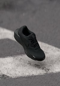 Chaussure de course noire avec le logo Nike, capturée en plein vol au-dessus d'un asphalte gris avec des marquages routiers blancs et quelques petits débris éparpillés.