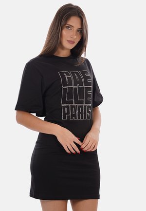 Femme aux longs cheveux bruns portant une robe noire à manches courtes avec le texte clouté « GHELLE PARIS » sur le devant.