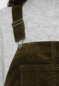 Lähi kuva tummanvihreistä cordura-haalareista, joissa on metallisolki, puettuna vaaleanharmaan ribbineuleisen poolopaidan päällä.