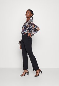 Blusa preta floral transparente com mangas longas, combinada com calças pretas ajustadas com fendas laterais e sandálias de salto alto pretas com tiras.