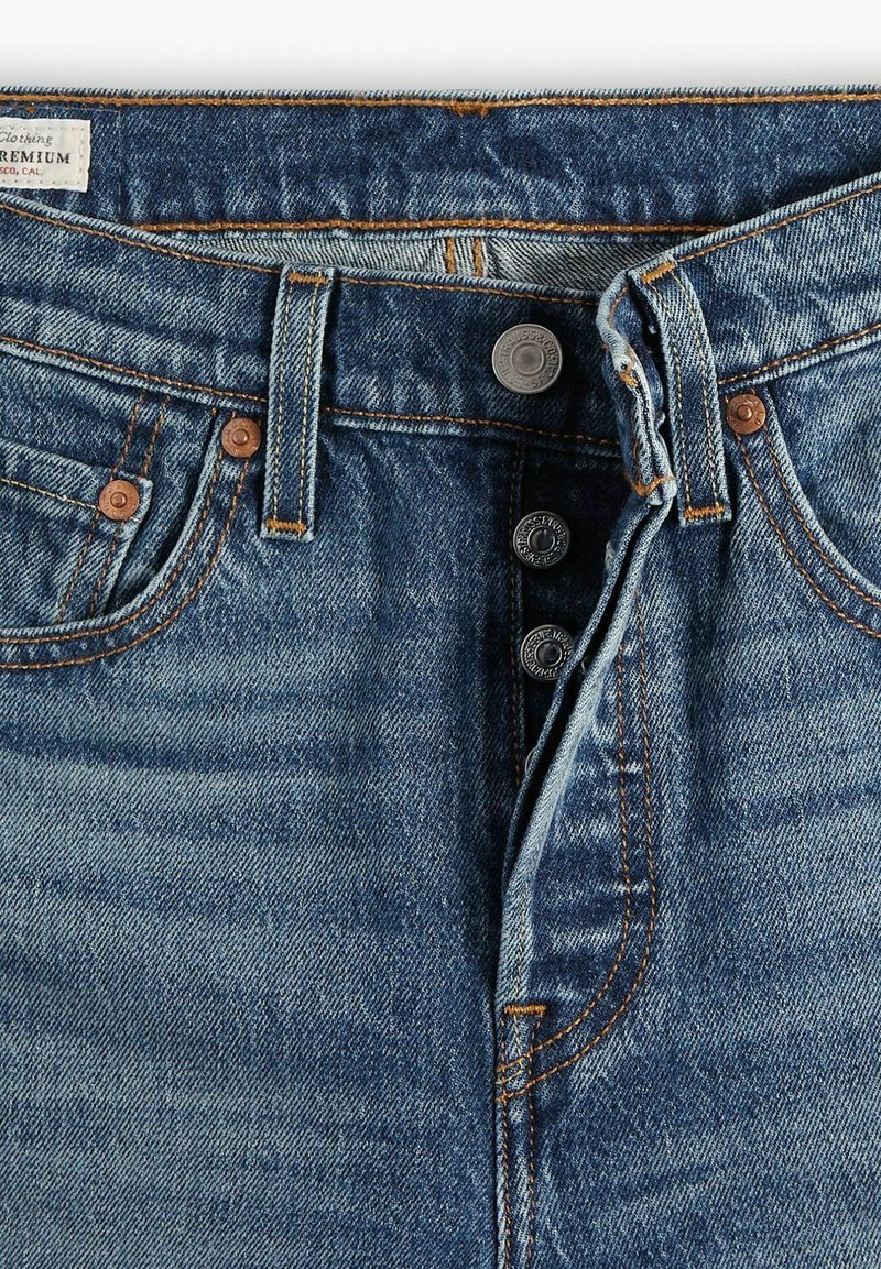Jean en denim dans un délavage bleu moyen, doté d'une fermeture à boutons, de rivets en cuivre et de coutures contrastantes orange le long des coutures.