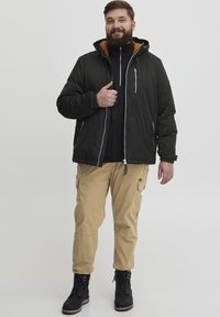 Sort isoleret jakke med lynlås-lommer og en brun foret hætte, parret med beige cargo-bukser og sorte snørebåndsstøvler.