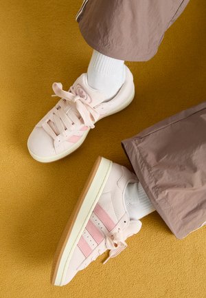 Roze suède sneakers met bijpassende strepen, witte veters en een crèmekleurige zool. Gecombineerd met witte sokken en losse bruine broeken op een gele achtergrond.