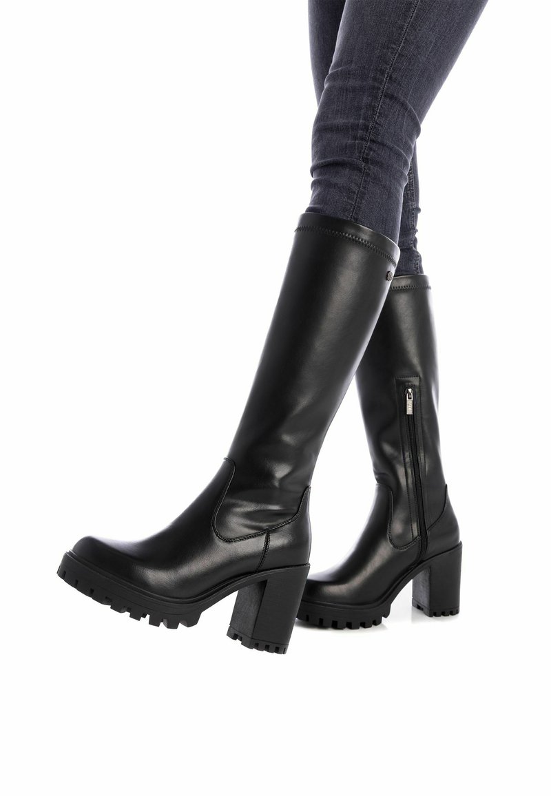 Botas de cuero negras hasta la rodilla con tacones gruesos, cremallera lateral y suela de goma texturizada. Acabado suave y detalles de costura visibles.