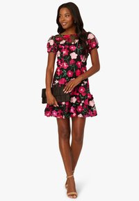 Květinové šaty s krátkými nadýchanými rukávy, černým základem, růžovými a červenými květy, přiléhavým živůtkem, rozšířeným lemem, doplněné černým psaníčkem a béžovými podpatky.