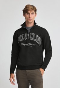 Svart fleece-tröja med halvdragkedja, med stor "POLO CLUB" text och etableringsdatum, i kombination med khakifärgade byxor.