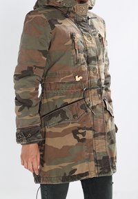 Kobieta w płaszczu z kapturem w wzór moro, z zamkami błyskawicznymi i kieszeniami zapinanymi na guziki, z regulowanym w pasie, a na piersi ma małą przypinkę w kształcie wiewiórki.