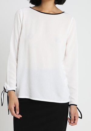 Blouse blanche à manches longues avec un col bateau, bord noir contrastant et liens ajustables aux poignets. Tissu léger et transparent.