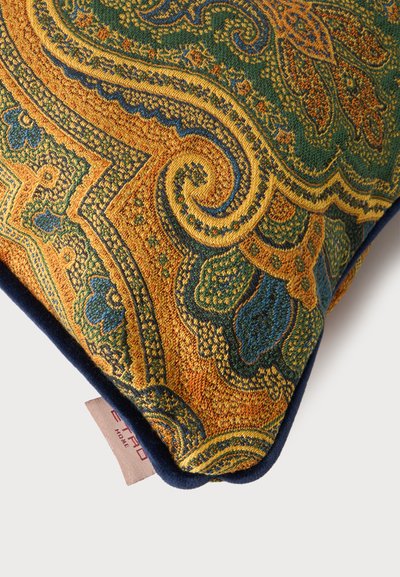 Coin de coussin décoratif avec un motif cachemire complexe dans des tons de vert, d'or et de bleu et une étiquette en tissu sur un fond uni.