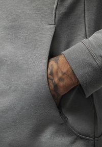 Felpa grigia con tasca. Tessuto texturizzato con una cucitura visibile. Una mano con un tatuaggio è appoggiata all'interno della tasca, mostrando il polsino della manica.