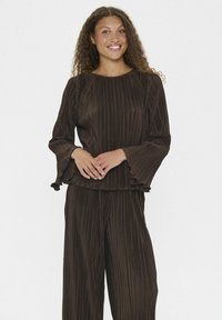Donna con capelli ricci che sorride, indossando una blusa lunga a maniche lunghe plissettata di un marrone scuro e pantaloni larghi abbinati, in piedi con le mani unite.