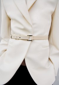 Vit blazer i ett mjukt tyg, med en figursydd midja och ett ljusbeige bälte med en metallspänne detaljer.