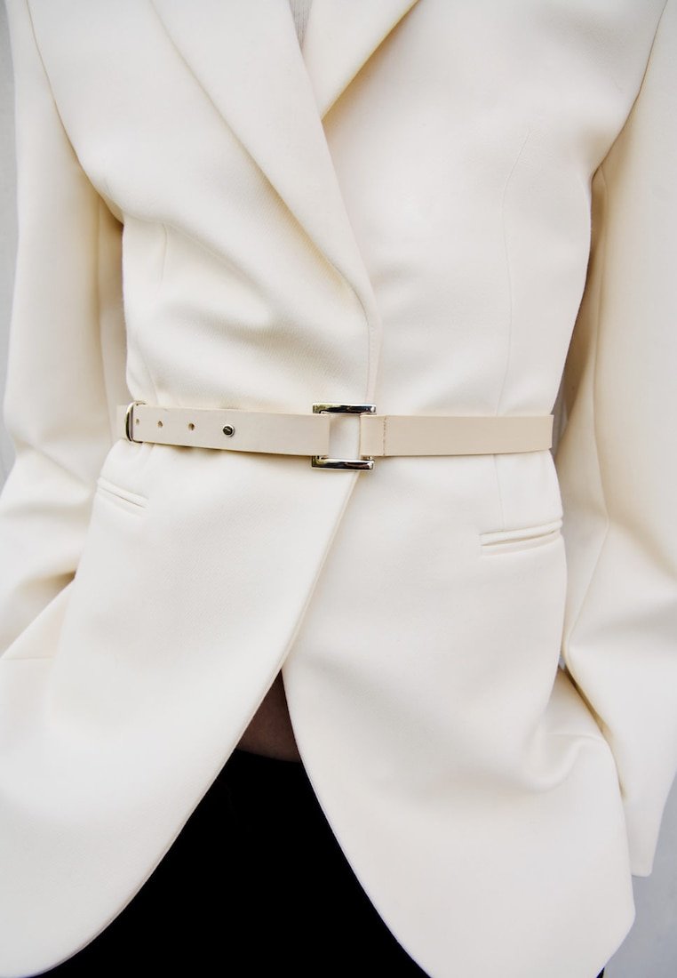 Vit blazer i ett mjukt tyg, med en figursydd midja och ett ljusbeige bälte med en metallspänne detaljer.
