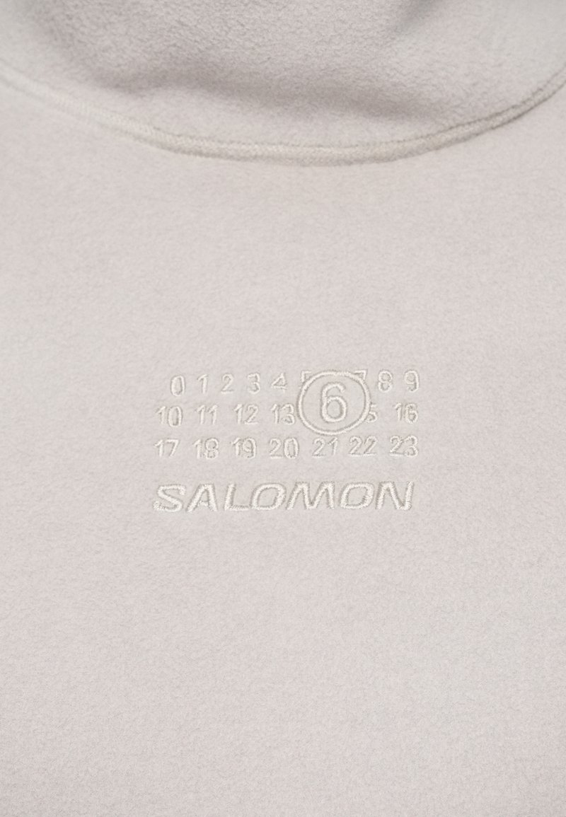 Close-up van lichtgrijze stof met geborduurde nummers 0-23 en het omcirkelde nummer 6 boven de merknaam SALOMON.