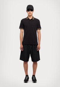 Čierne polo tričko s vzorovaným golierom, čierne šortky a čierne tenisky. Doplnky zahŕňajú čiernu čiapku a tmavé slnečné okuliare.