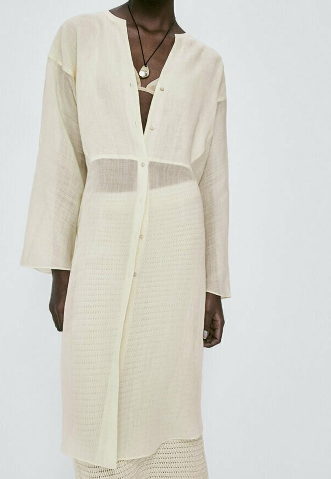 Robe longue beige clair semi-transparente boutonnée avec des manches larges portée sur un pantalon en crochet assorti et un collier visible.