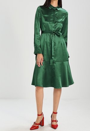 Femme portant une robe-chemise verte brillante boutonnée avec des manches longues et une taille nouée, associée à des chaussures à talons rouges avec des brides à la cheville.