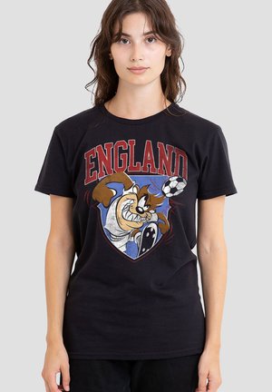 Junge Frau trägt ein schwarzes T-Shirt mit dem Wort England und einem Cartoon-Tasmanischen Teufel, der vorne einen Fußball kickt.