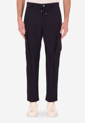 Uomo che indossa pantaloni cargo neri con coulisse e tasche a pattina, abbinati a sneakers off-white, in piedi su uno sfondo bianco.