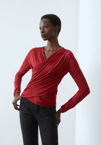 Red long-sleeve top with a wrap design and draped texture. Paired with black jeans, showcasing a fitted silhouette against a neutral background.