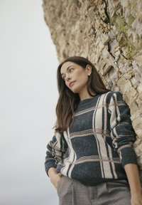 Strickpullover in Dunkelblau mit beigem Karomuster und gerippten Bündchen, kombiniert mit hellgrauen Hosen, vor einer strukturierten Steinwand.