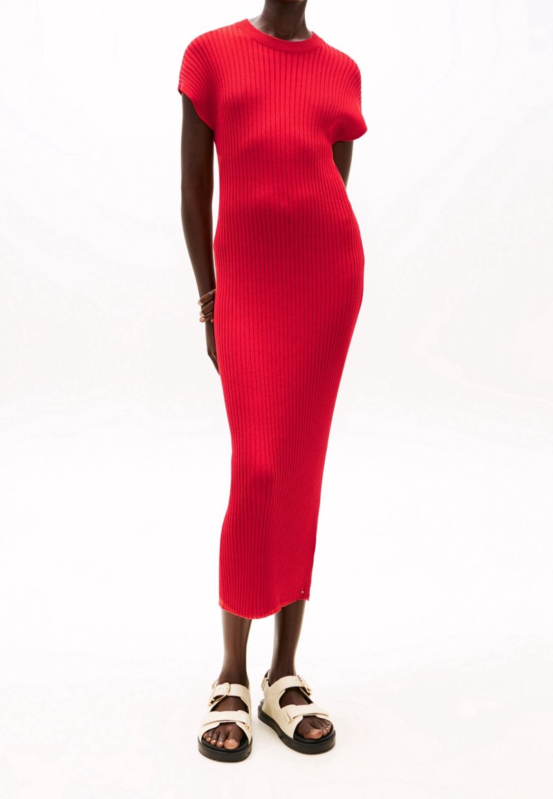 Personne portant une robe midi rouge ajustée à côtes avec manches courtes et des sandales blanches à boucle sur fond blanc.