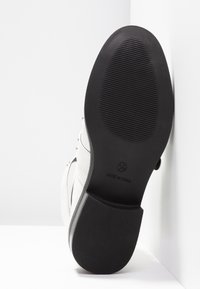 Semelle de chaussure blanche montrant un dessous noir texturé de taille 4 1/3 et texte "Made in China", inclinée contre un mur d'angle blanc.
