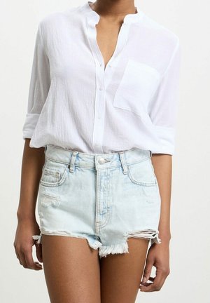 Vrouw die een wit, lang mouwen, overknopte blouse met opgerolde mouwen draagt en lichtblauwe versleten denimshorts met rafelige zomen.