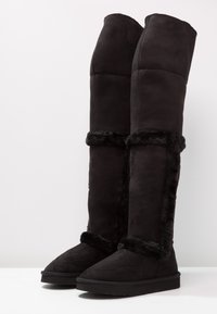 Svarta knähöga stövlar i mjukt mocka med en plysch av syntetisk päls längs sidorna och toppen. Platt sula och rund tådesign.