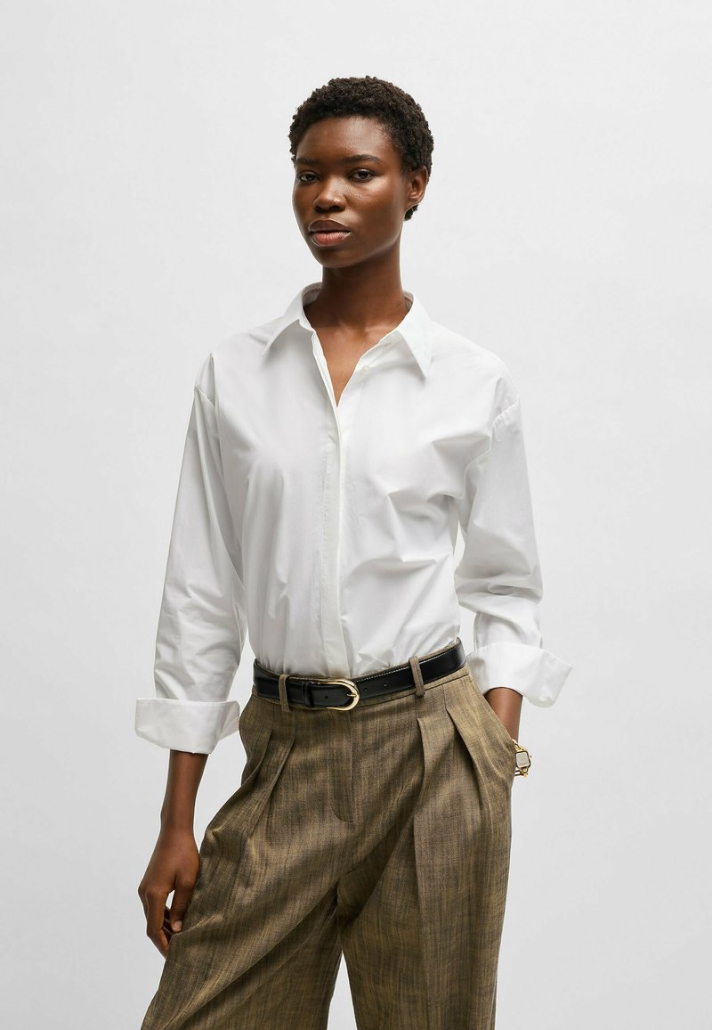 Camisa blanca de manga larga con botones, de corte relajado y con los puños remangados, combinada con pantalones beige de patrón y un cinturón negro.