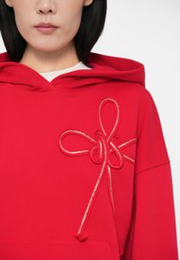Γυναίκα που φοράει κόκκινο hoodie με ανάγλυφες λεπτομέρειες σε σχήμα κορδέλας και μεταλλικές χρυσές λεπτομέρειες στο στήθος, σε άσπρο φόντο.