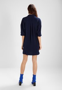 Donna con lunghi capelli castani, che indossa un vestito blu navy largo e stivaletti alla caviglia blu brillante, in piedi con la schiena rivolta su uno sfondo chiaro e uniforme.
