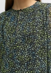 Blumenbluse mit schwarzem Hintergrund und kleinen blauen und grünen Blumen, runder Ausschnitt mit Knopfverschluss und strukturiertem Stoff.