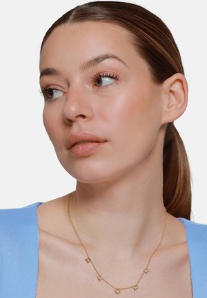 Donna con capelli castani legati in una coda bassa, indossa una maglietta blu e una delicata collana d'oro con piccoli ciondoli a forma di farfalla, guarda verso sinistra.