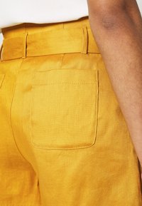 Gros plan d'une personne portant un pantalon jaune vif avec une poche arrière et des passants pour ceinture, montrant une partie de son bras et un haut clair.