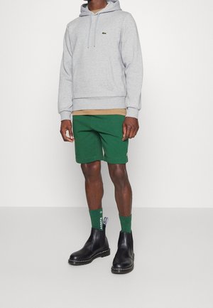 Sweat à capuche gris avec une poche avant, superposé sur une chemise beige. Shorts verts, bottines Chelsea noires et chaussettes vertes avec un logo blanc.