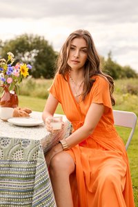 Femme en robe orange assise à l'extérieur à une table avec un verre à la main, des fleurs dans un vase et des croissants sur une assiette à proximité.
