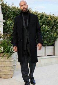 Abrigo de lana negro con un corte entallado, combinado con un suéter de cuello alto oscuro y pantalones de rayas, complementado con elegantes zapatos negros.