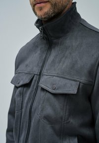 Chaqueta gris con cremallera hecha de tela suave, que cuenta con dos bolsillos con tapa y botones a presión, un cuello alto y una textura suave.