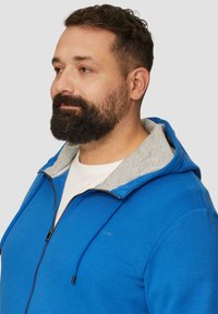 Niebieska bluza z kapturem na zamek błyskawiczny z szarym wykończeniem, posiadająca sznurek do regulacji kaptura, zamek z przodu oraz minimalny detal logo na klatce piersiowej. Materiał bawełniany.