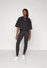 Czarny krótki t-shirt z logo i czarne legginsy z małym detalem z logo. Modelka nosi białe buty sportowe. Gładka, elastyczna tkanina.