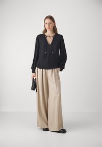 Black blouse with a V-neck and tassel details, paired with high-waisted beige wide-leg trousers. Model holds a small black handbag.