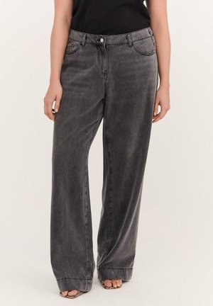 Kvinde iført løstsiddende, vaskede sorte jeans med synlige syninger, forlommer og knaplukning, kombineret med åbne sandaler.