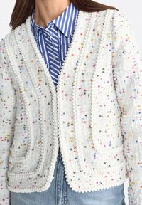 Weiße strukturierte Jacke mit mehrfarbigen Sprenkeln und Pomponbesatz, getragen über einem blau-weiß gestreiften Hemd mit Kragen und hellblauen Jeans.